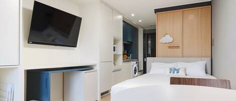 A modern hotel room with a flat-screen TV mounted on the wall, a compact kitchenette, a bed with a wooden headboard, and a ceiling-mounted light fixture.