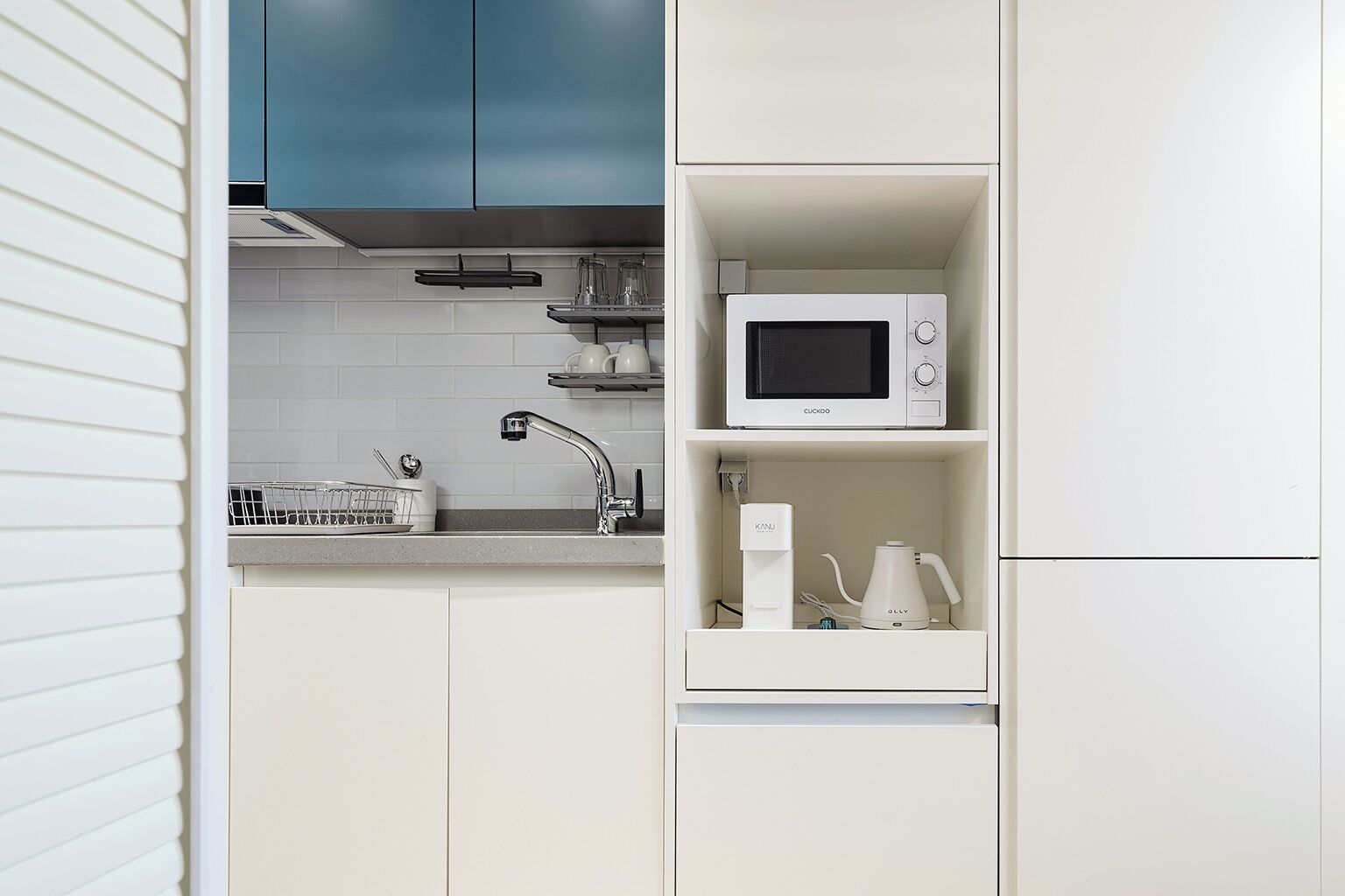 A compact kitchen with a microwave, kettle, and a dish rack.
