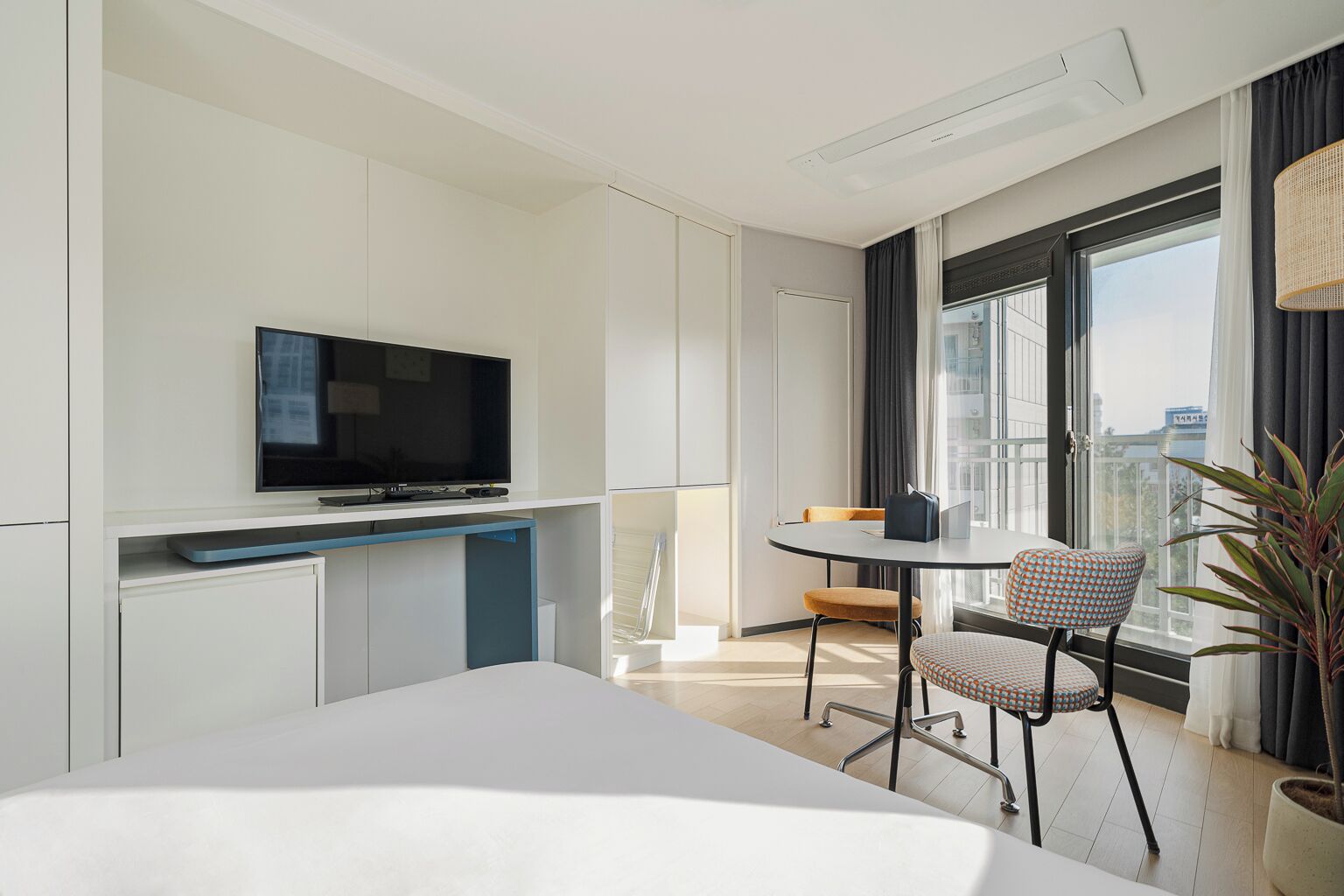Habitació d'hotel moderna amb televisió de pantalla plana, una taula de dinar amb dues cadires, una planta en test i un gran finestral amb vista a la ciutat.