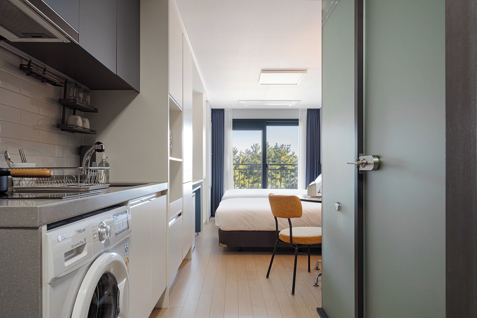 A compact hotel room with a kitchenette, a bed, a chair, and a view of the outdoors.