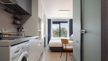 A compact hotel room with a kitchenette, a bed, a chair, and a view of the outdoors.