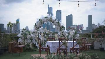 Et udendørs opholdsområde med et bord og stole, omgivet af hvide blomster og grønt, med en bysilhuet i baggrunden.