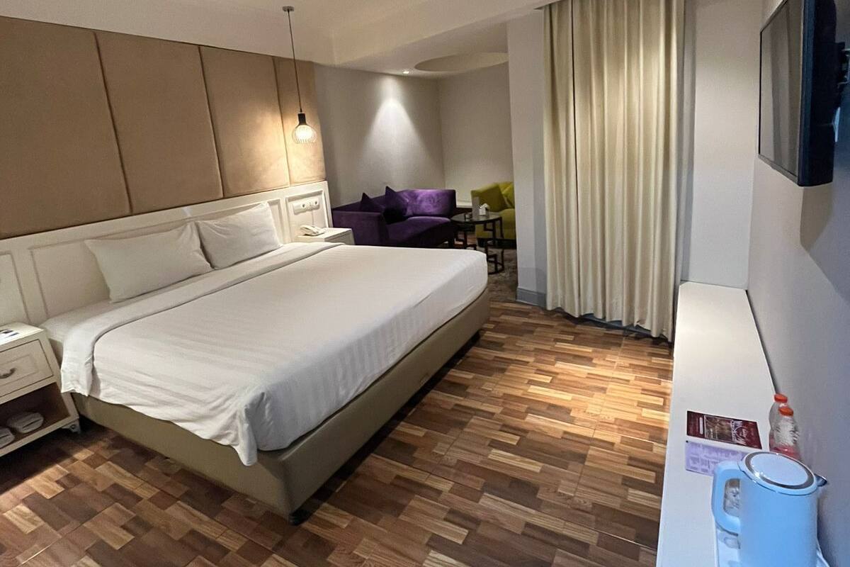 A modern hotel room with a large bed, a desk with a kettle, and a TV on the wall.