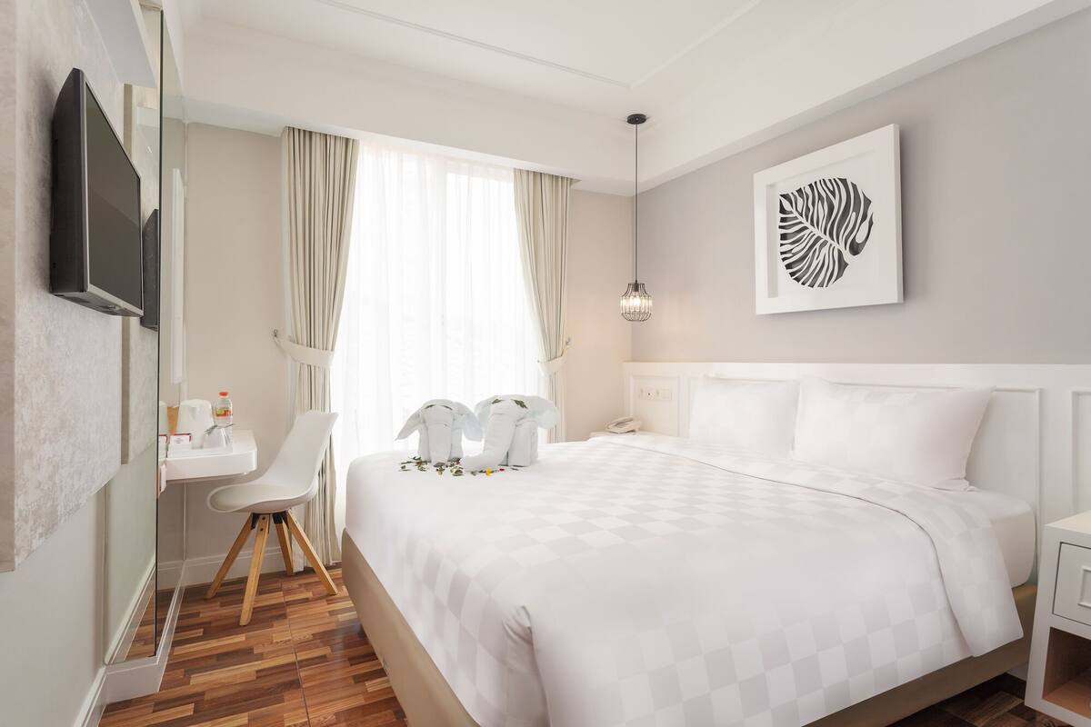 A hotel room with a large bed, a desk, a chair, a bedside table, a telephone, and a wall-mounted TV.