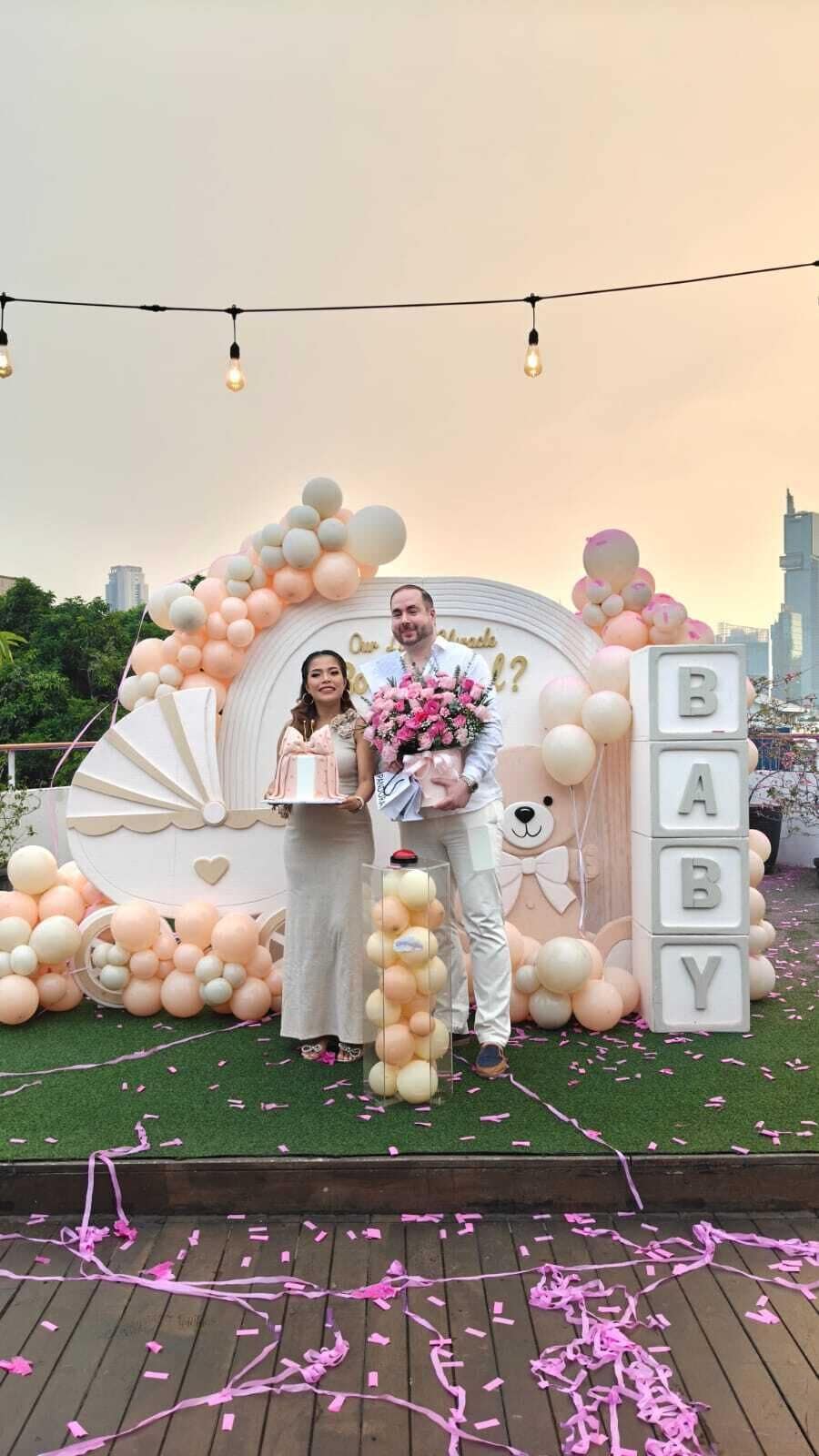 A couple standing in front of a baby shower setup with balloons, a fan, and a 'BABY' sign.