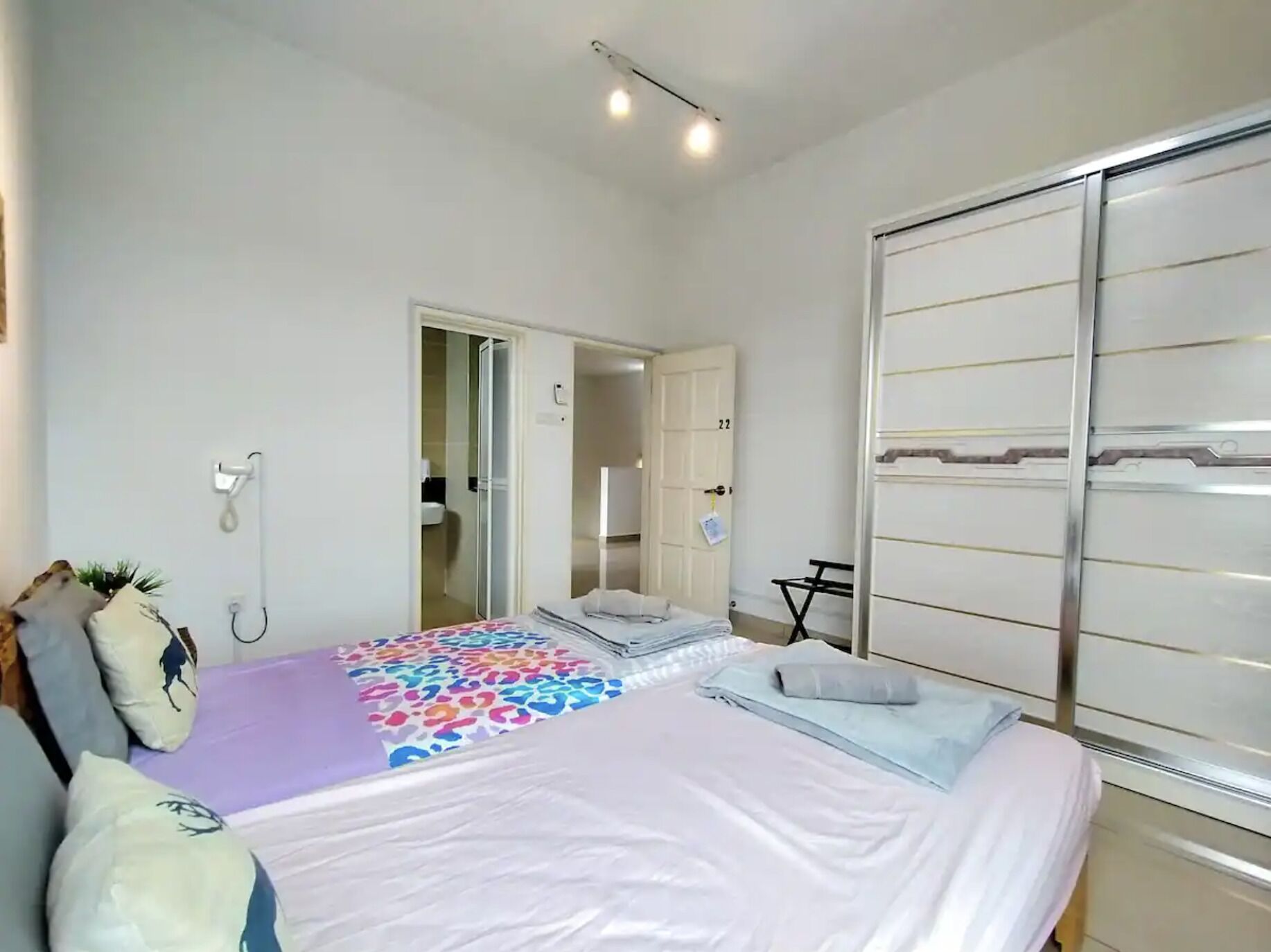 Une chambre à coucher avec un lit, une armoire, une salle de bain visible à travers une porte ouverte, et une petite table avec une chaise.