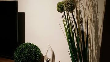 A room with a wooden shelf holding a television, a decorative plant, and a white vase with a spherical object.