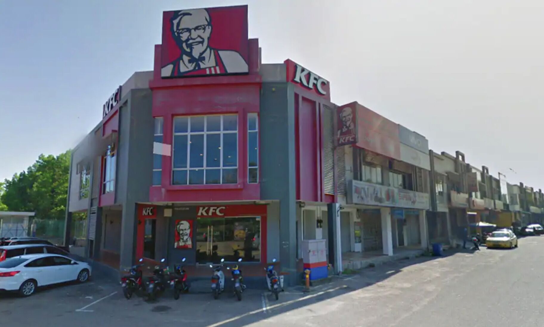 En KFC restaurant med et stort rødt og hvidt skilt, flere motorcykler foran og en række butikker langs gaden.