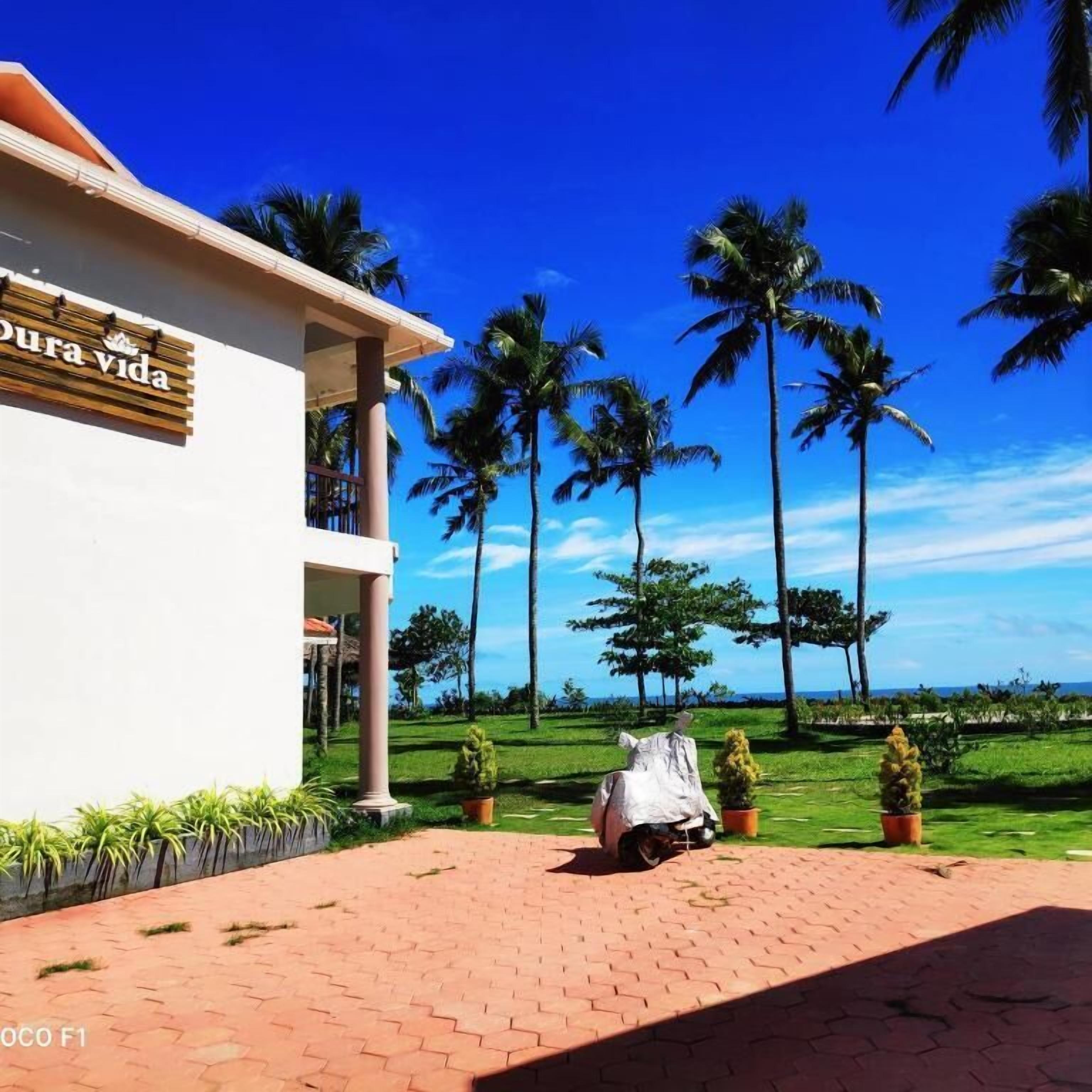 A hotel with a sign reading "Dura Vida" and a paved entrance area with palm trees and a view of the ocean.