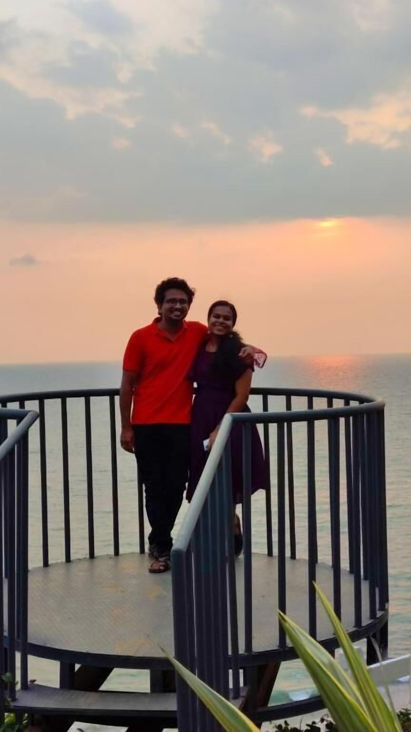 Una pareja de pie en un balcón circular con barandilla, contemplando un cuerpo de agua al atardecer.