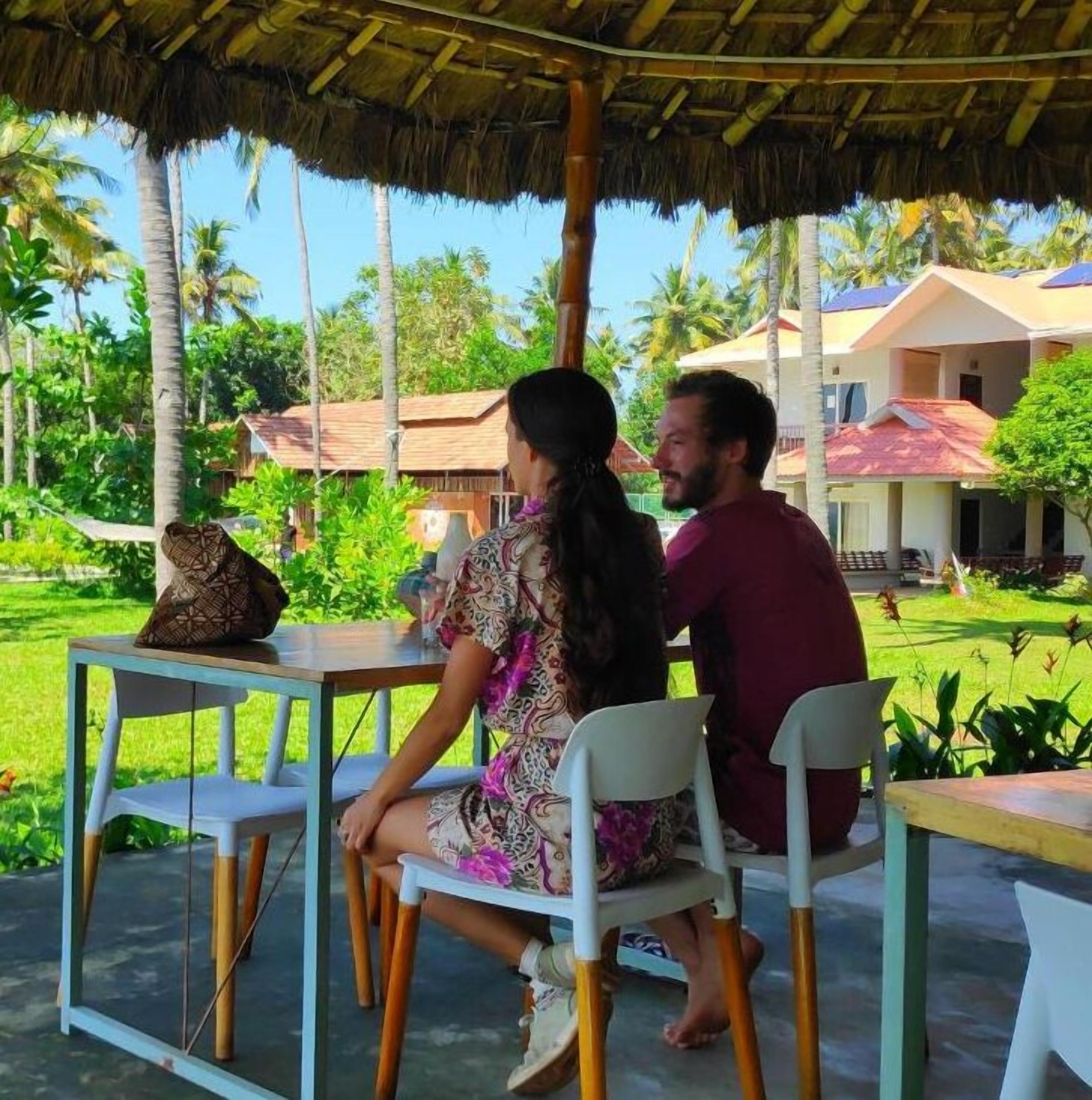 Una pareja sentada a una mesa bajo un techo de paja, rodeada de abundante vegetación y palmeras.