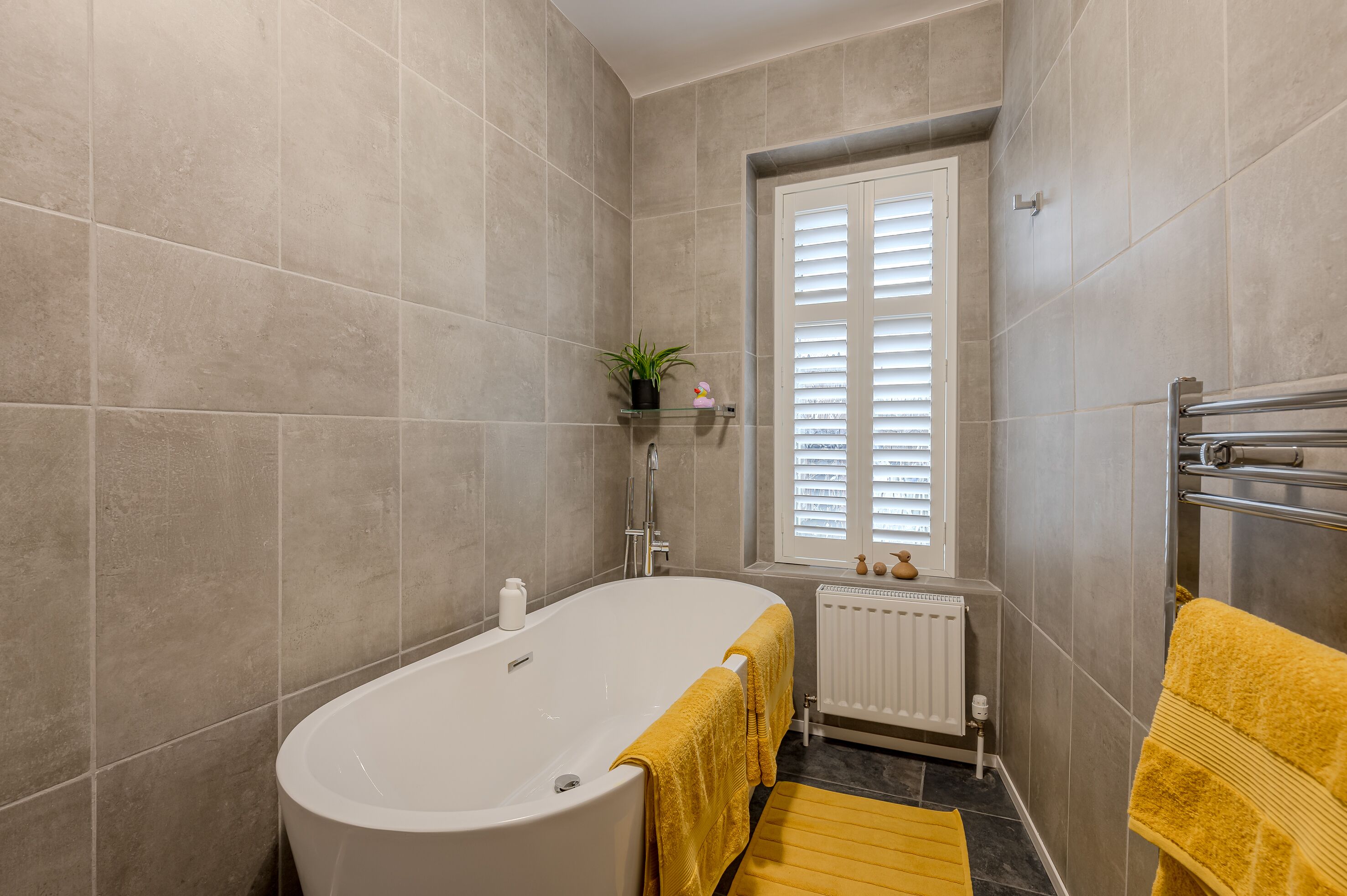 A bathroom with a bathtub, a window with blinds, a radiator, and a yellow towel on the floor.