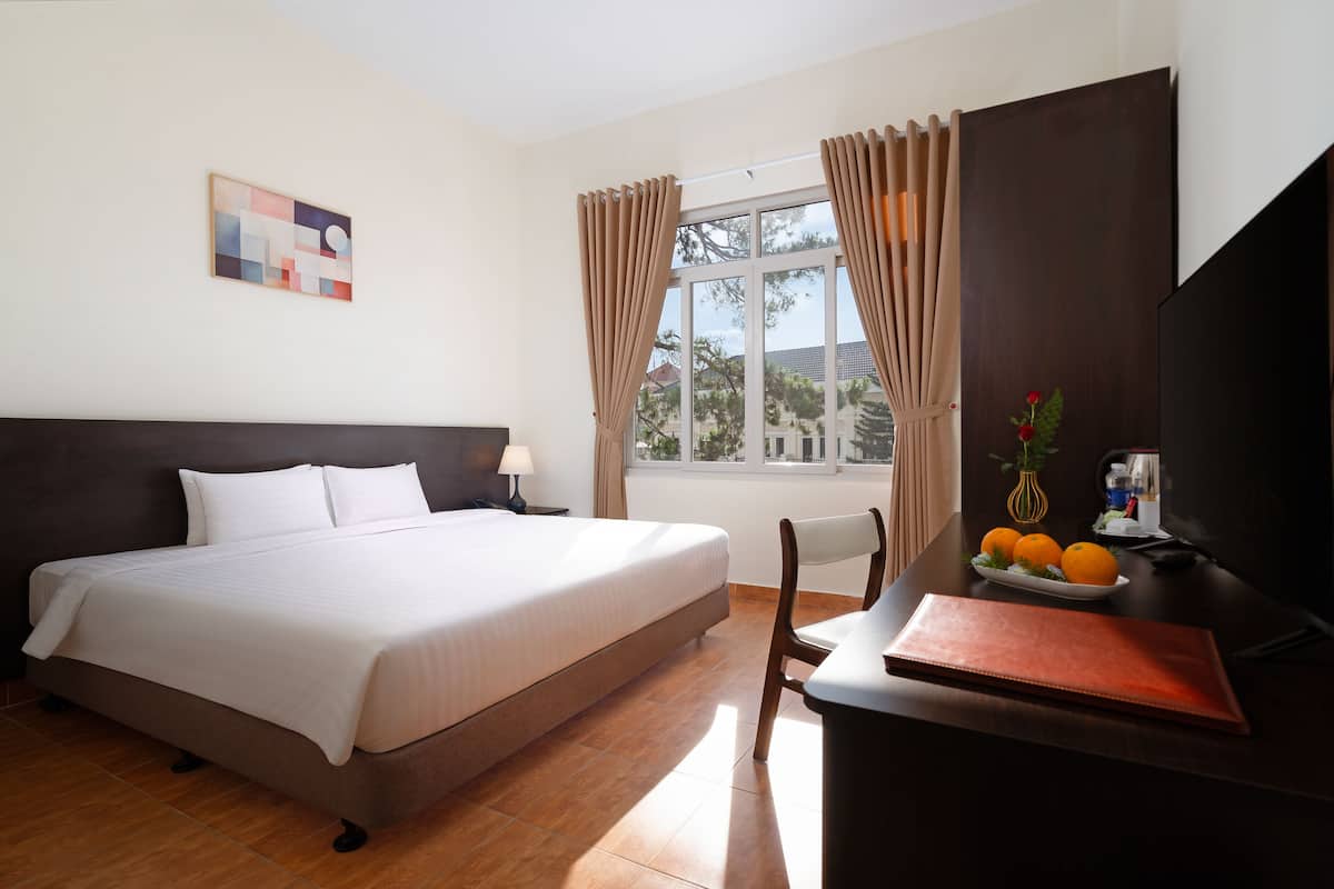 A hotel room with a large bed, a desk with a laptop, a vase with flowers, and a window with curtains.