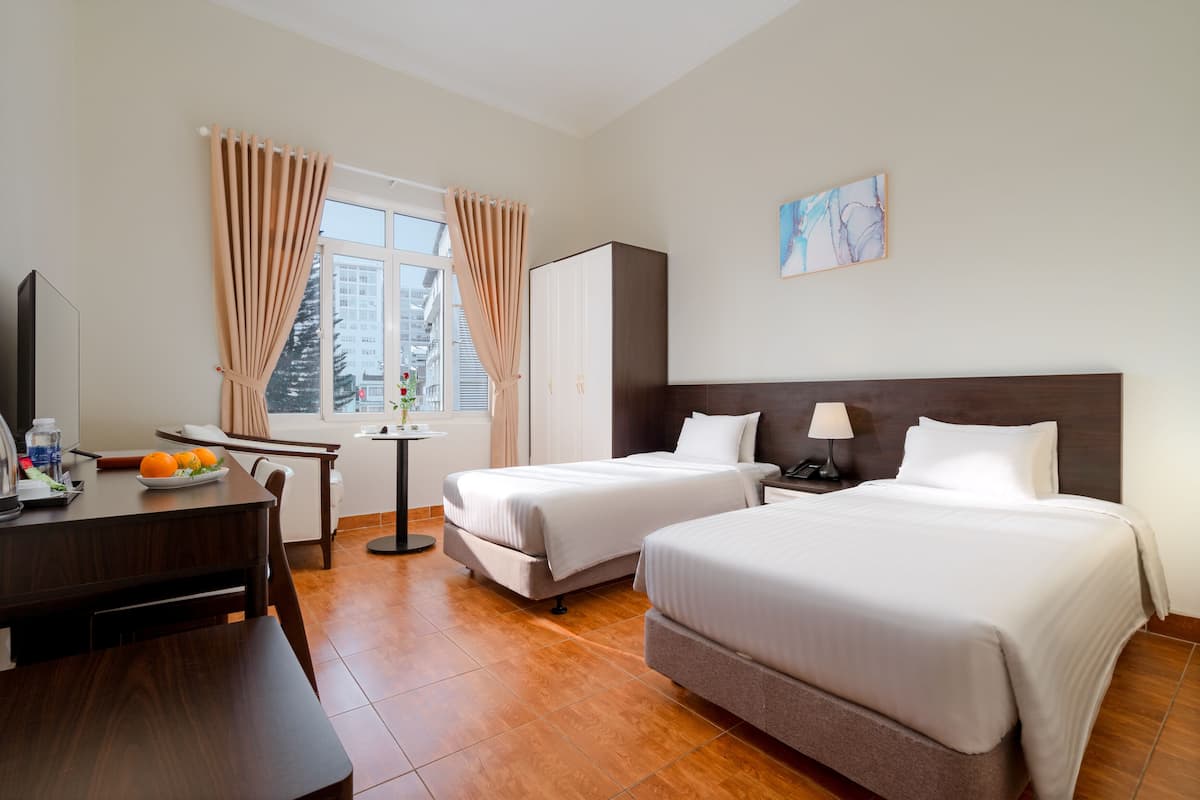 A hotel room with two beds, a desk with a chair, a small table, and a window with curtains.
