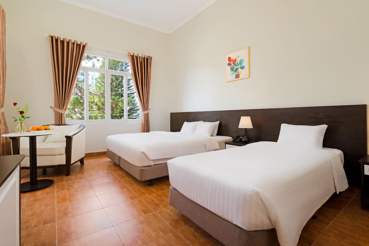 A hotel room with two beds, a desk, a chair, a window with curtains, and a painting on the wall.