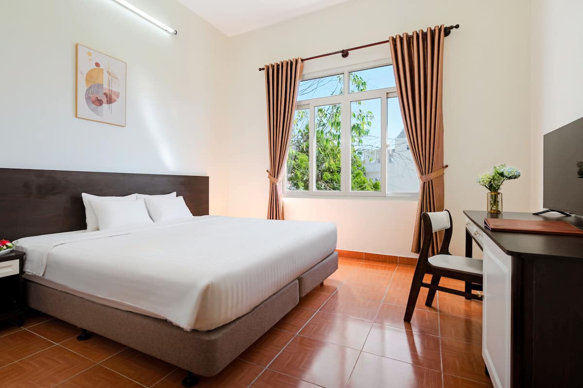 A hotel room with a bed, a desk, a chair, a TV, a window with curtains, a lamp, and a framed picture on the wall.