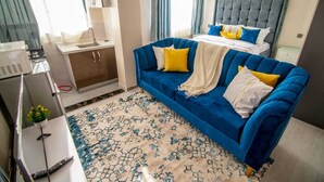 Ein Hotelzimmer mit einem blauen Sofa, einem Bett mit grauem Kopfteil, einem kleinen Schreibtisch und einem Fenster mit Vorhängen.