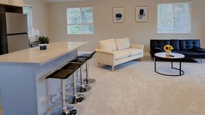 Eine moderne Küche mit Barbereich, einem beigen Sofa, einem schwarzen Sofa und einem runden Couchtisch mit einer Vase voller Sonnenblumen.