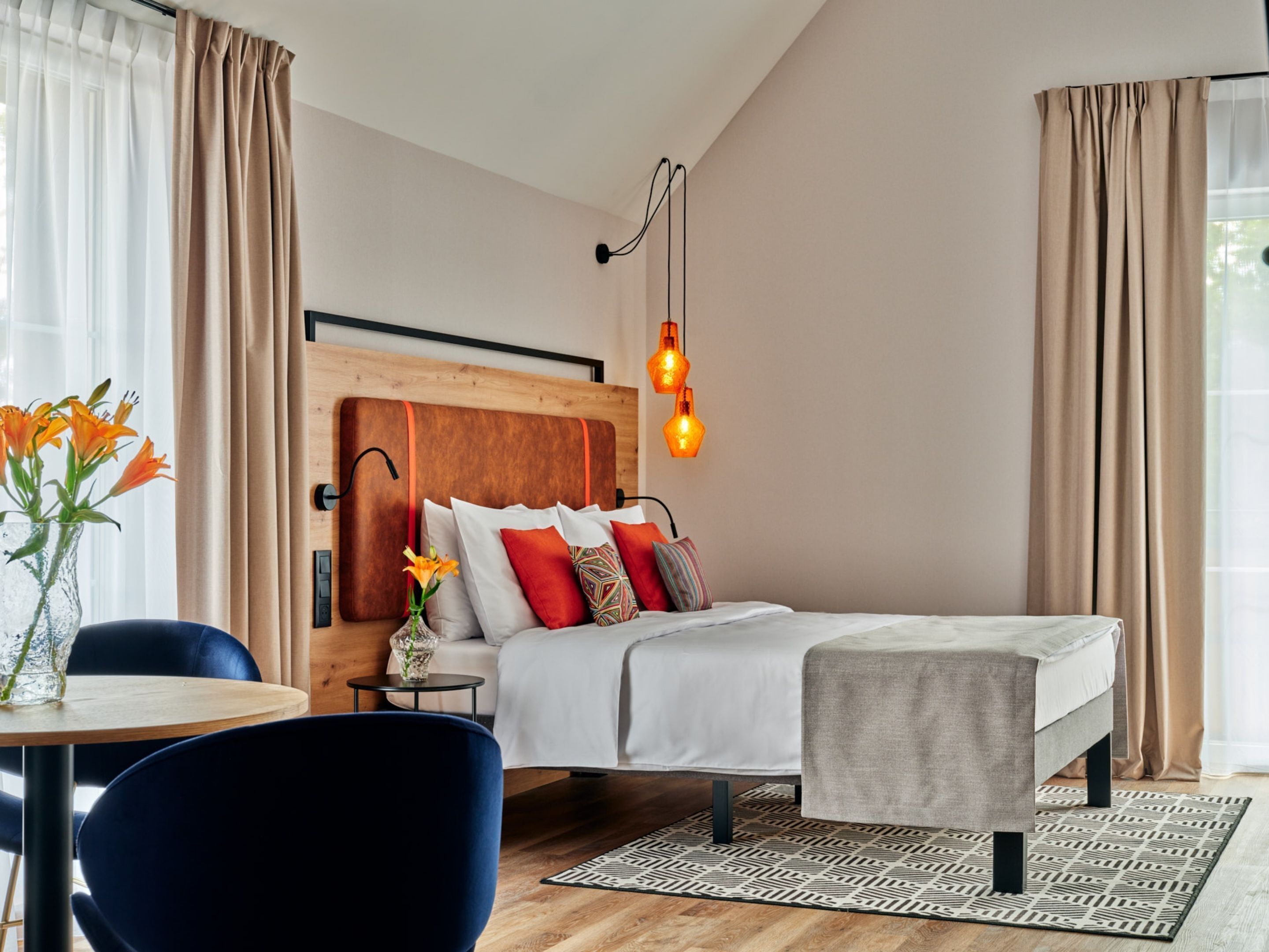 A modern hotel room with a wooden headboard, a bed with white bedding, a small table with a vase of flowers, and a blue chair.