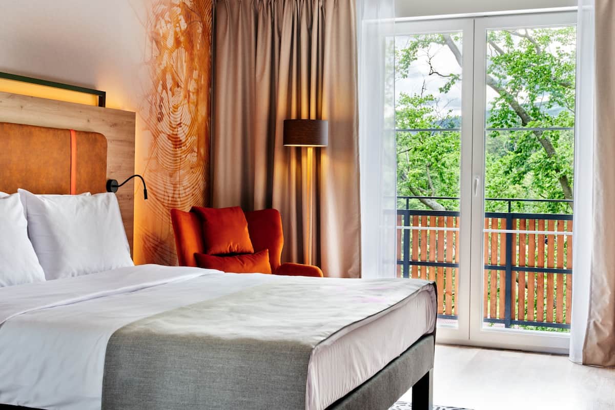 A hotel room with a bed, a chair, a large window, and a view of greenery outside.