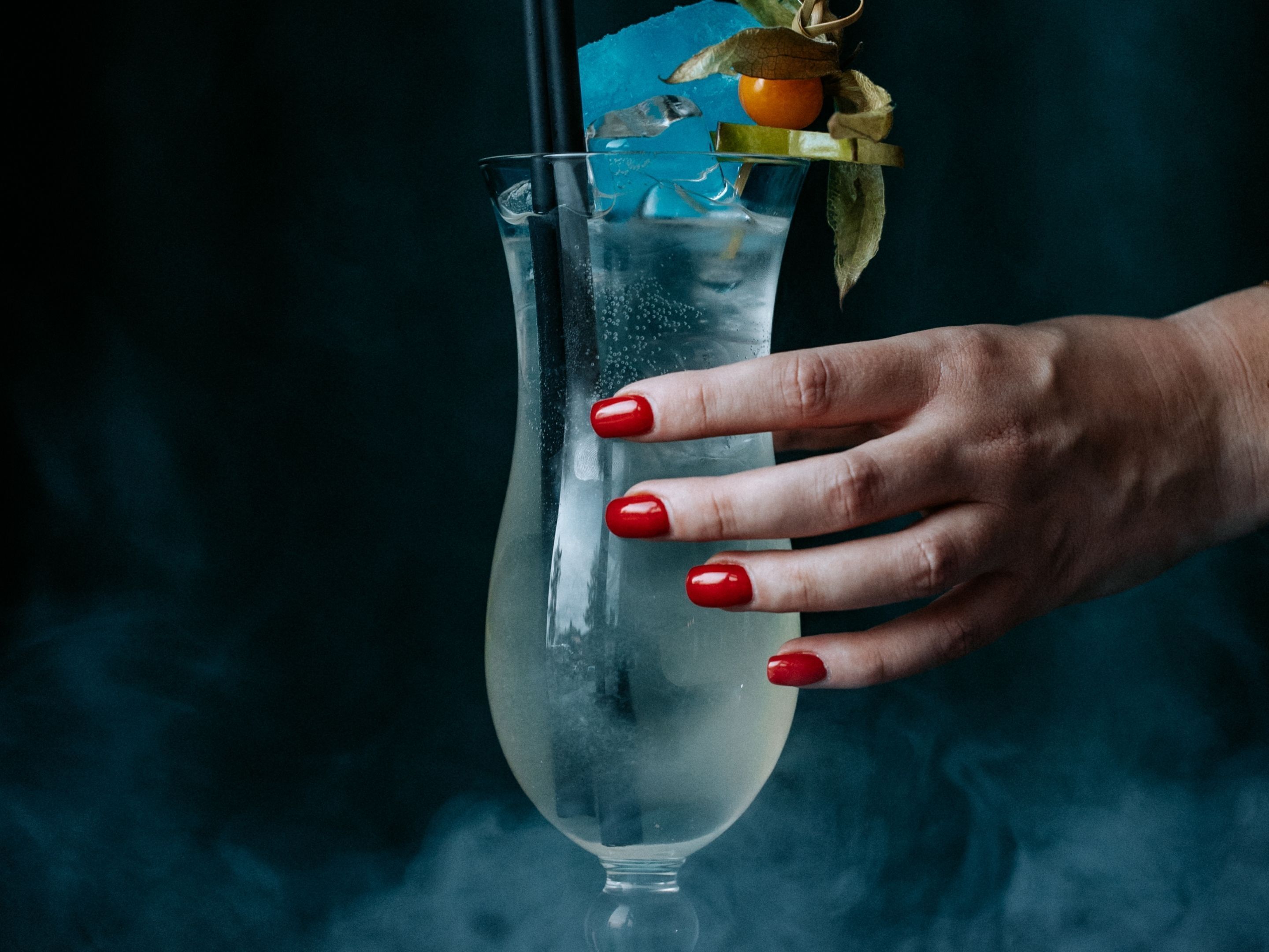 Una mano sosteniendo un vaso alto con un líquido transparente y hielo, adornado con fruta y una hoja.