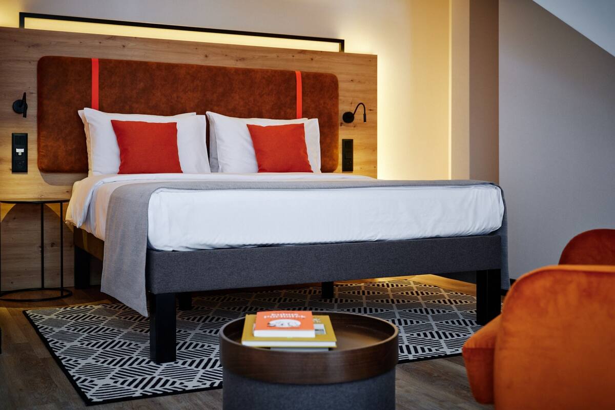 A modern bedroom with a wooden headboard, a patterned rug, a bedside table with a book, and an orange chair.
