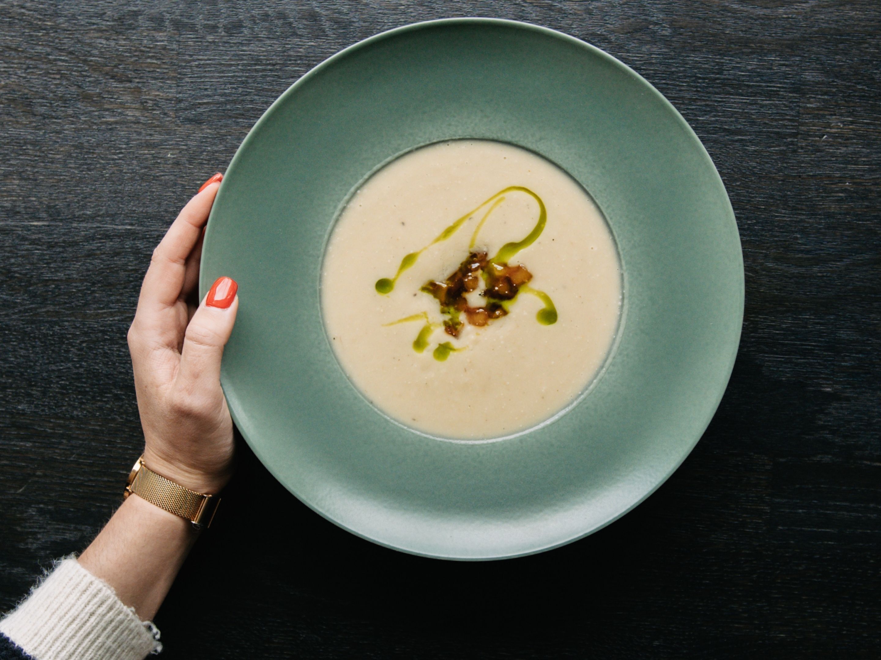 A hand holding a plate with a creamy soup garnished with green leaves and a drizzle of sauce.