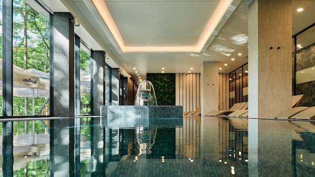 A modern indoor swimming pool with clear water, a central fountain, and a view of trees outside. - Hotel Bergo Resort & SPA (Szklarska Poreba)