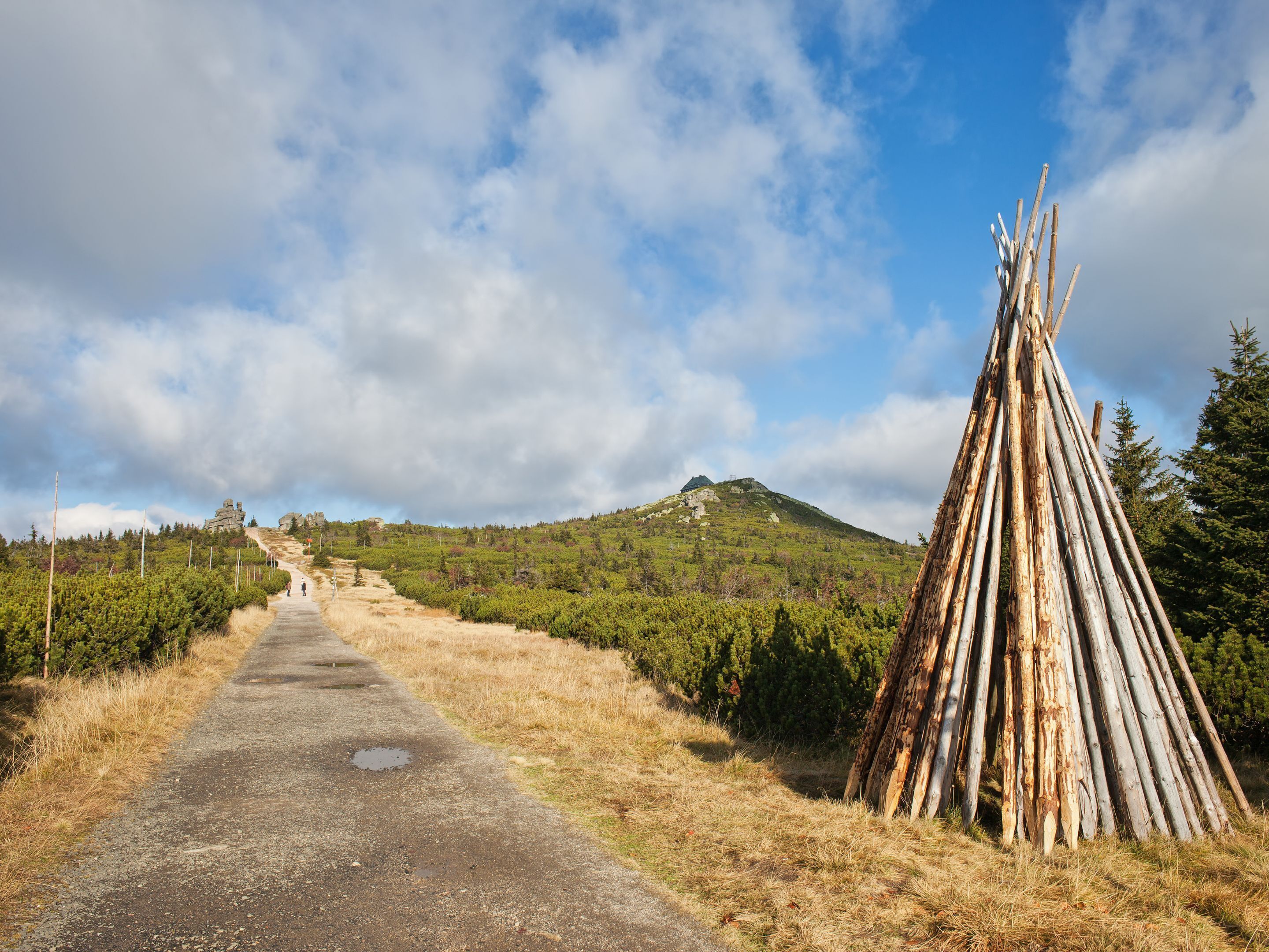 Ein schmaler Feldweg führt zu einer Holzhütte in einer natürlichen Umgebung, im Hintergrund ist eine Berglandschaft zu sehen.