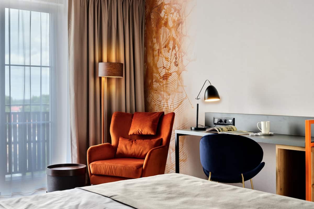 A modern hotel room with a large bed, a desk, a chair, a lamp, and a view of the outdoors.