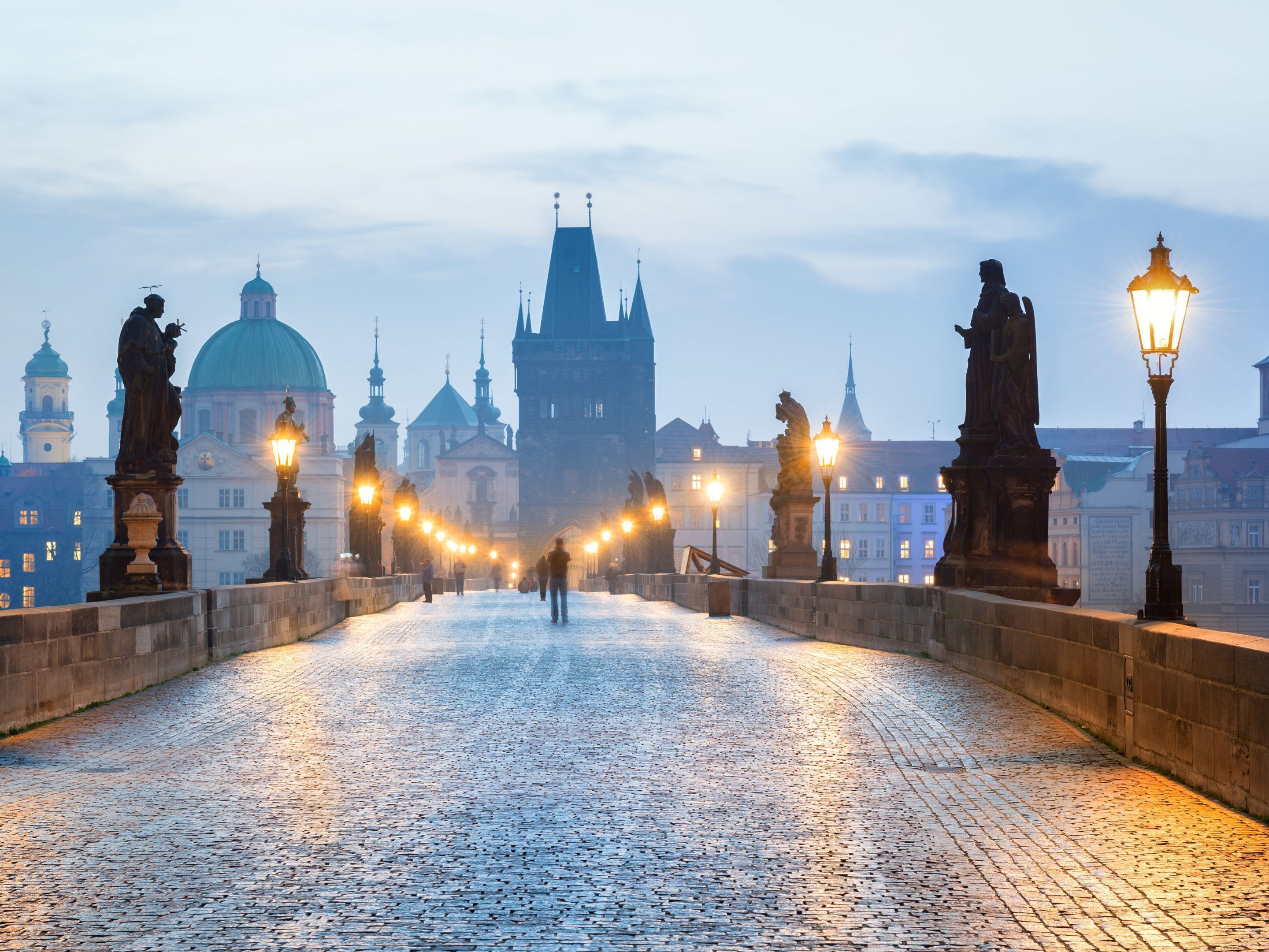 A cobblestone bridge with statues and street lamps, leading to a historic building with domed architecture.