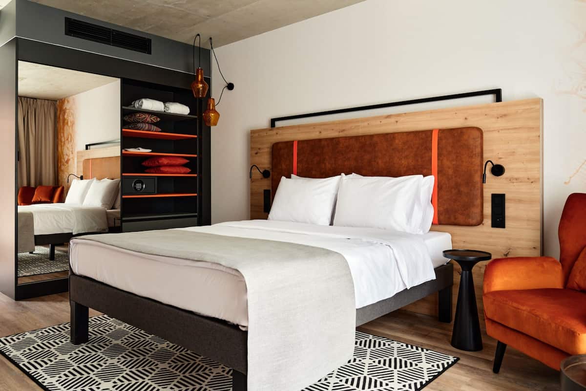 A modern bedroom with a large bed, a wooden headboard, a black side table, a red armchair, and a built-in wardrobe with shelves.