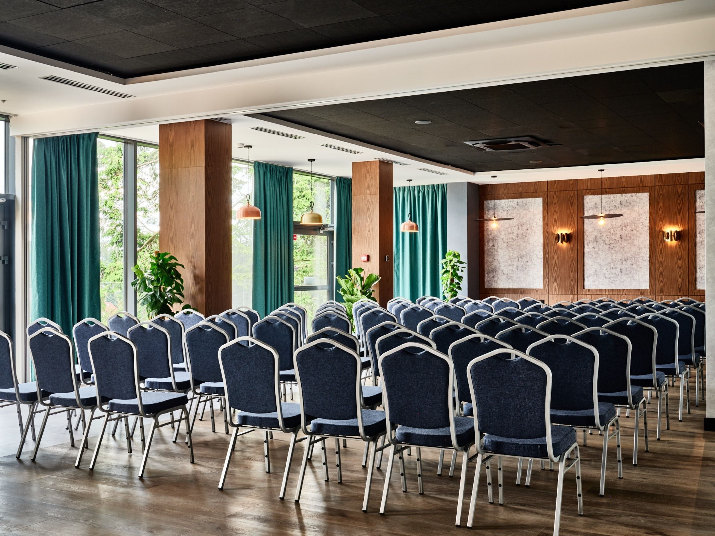Une salle de conférence avec des rangées de chaises, de grandes fenêtres avec des rideaux, des panneaux de bois et des luminaires suspendus.