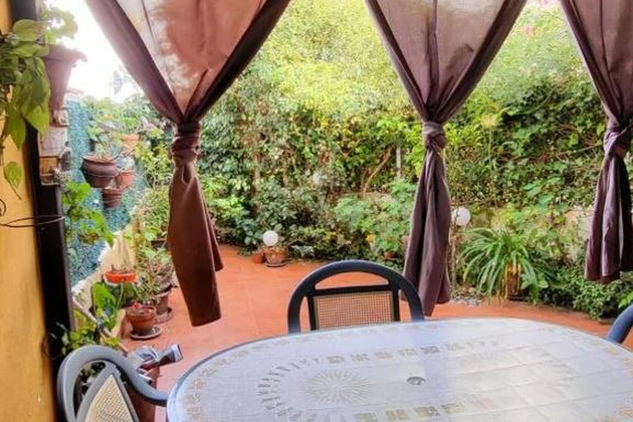 A round table with a patterned tablecloth, surrounded by chairs, in a garden area with potted plants and curtains.