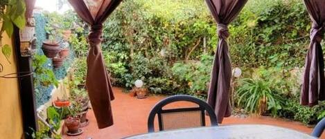 A round table with a patterned tablecloth, surrounded by chairs, in a garden area with potted plants and curtains.