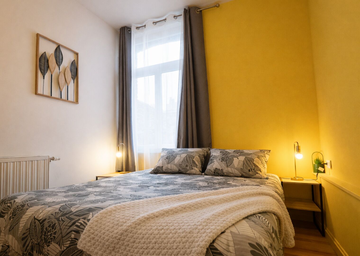 A bedroom with a bed, a nightstand, a lamp, a radiator, and a window with curtains.