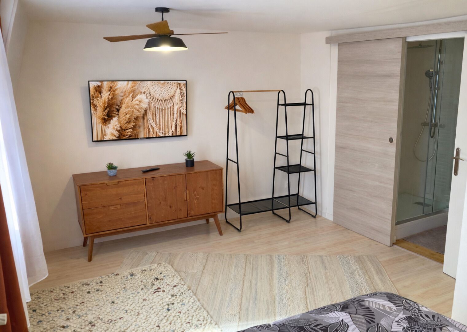 A room with a wooden cabinet, a black metal shelf, a ceiling fan, a wall-mounted TV, and a glass shower enclosure.
