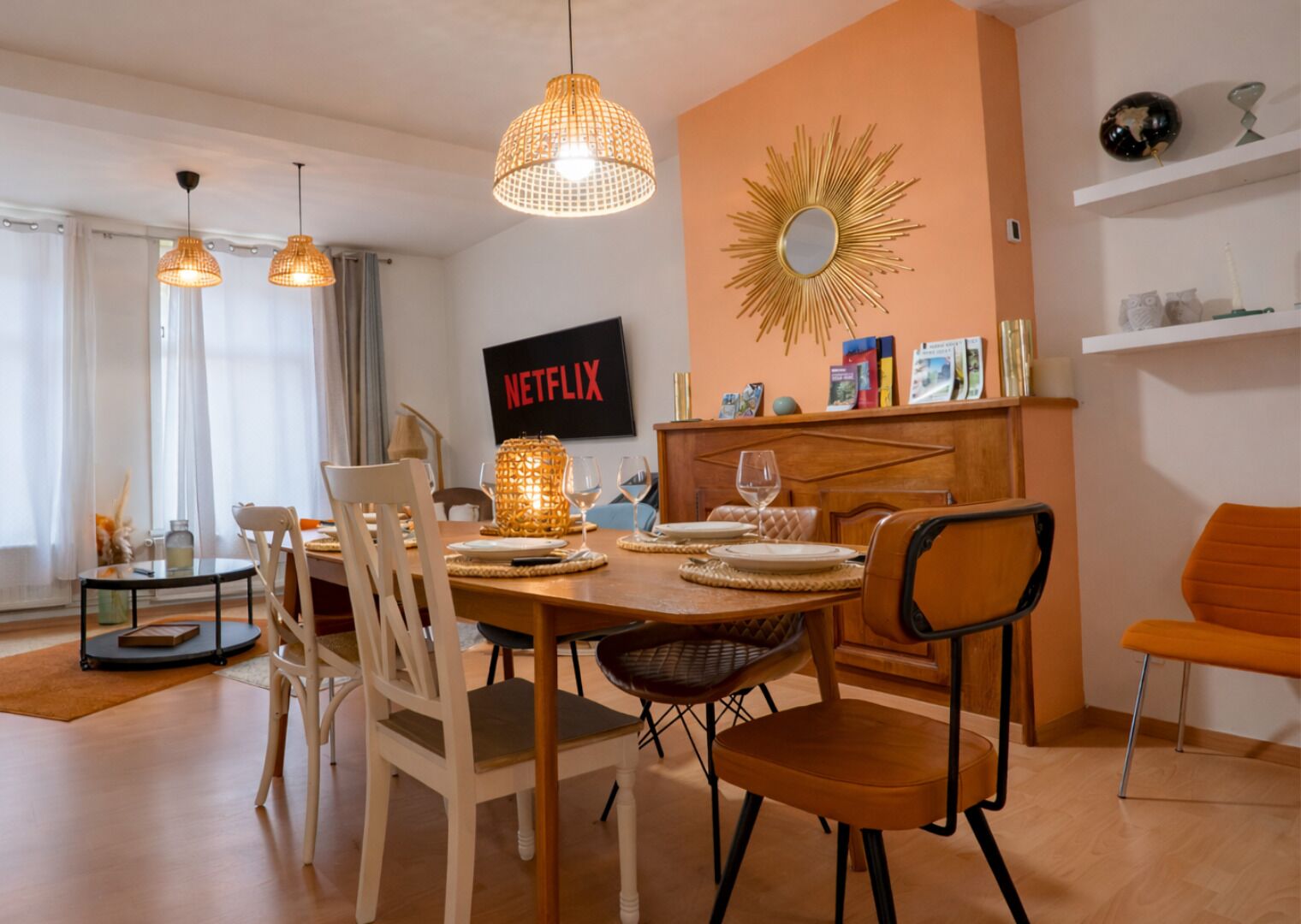 A dining area with a table set for four, chairs, pendant lights, a wall-mounted TV, and a decorative mirror.