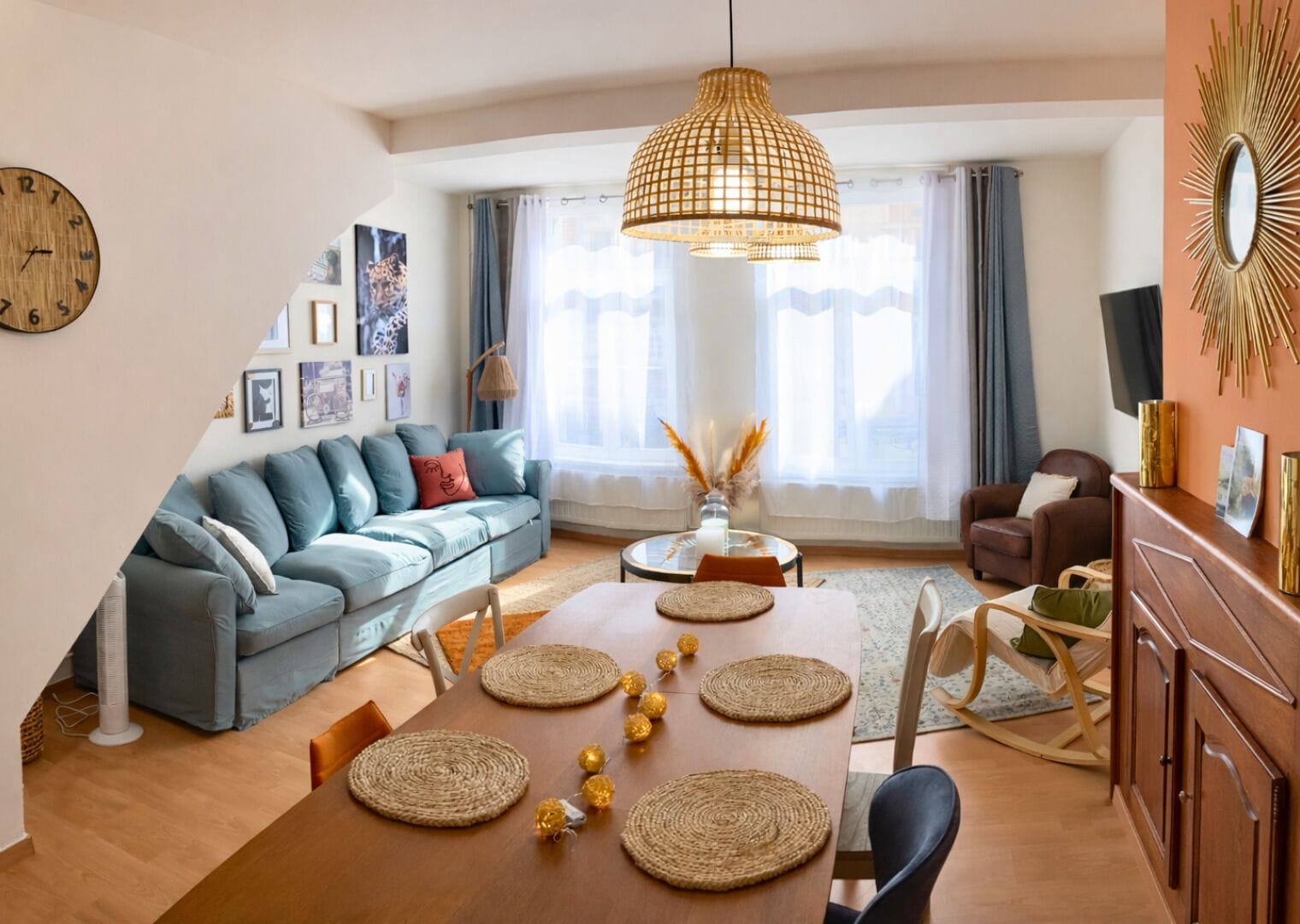 A modern living room with a sofa, dining table, and chairs, a hanging light fixture, and a clock on the wall.