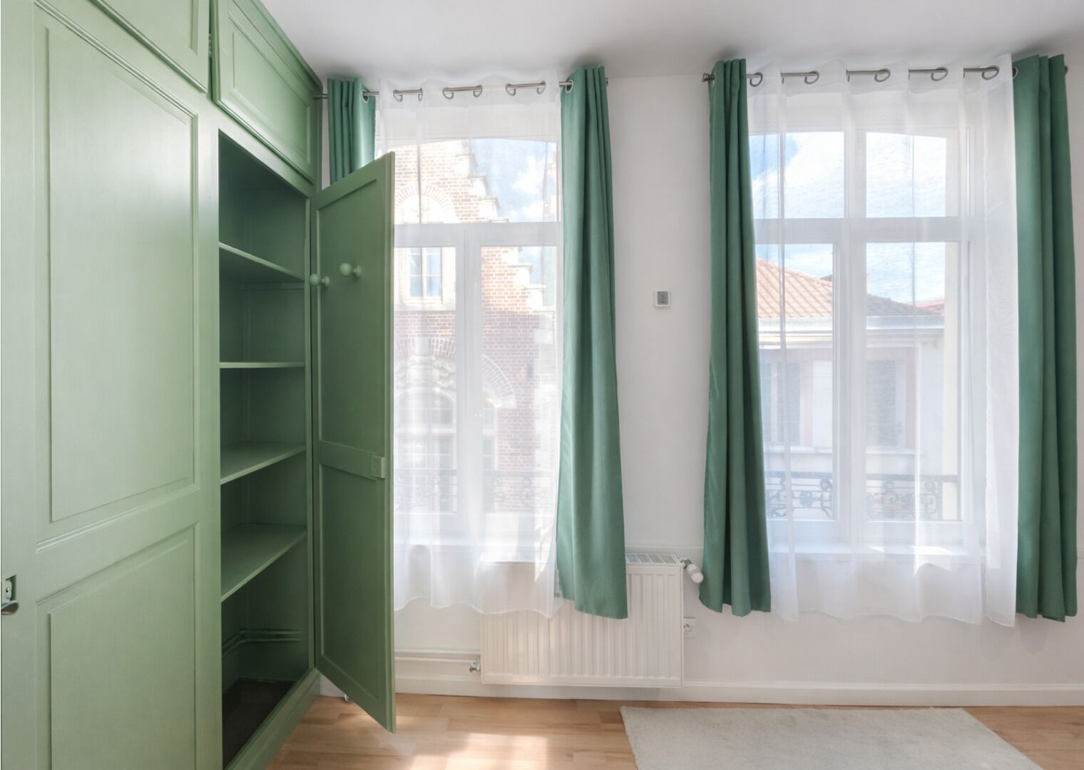 A room with a green wardrobe, a radiator, and a window with green curtains.