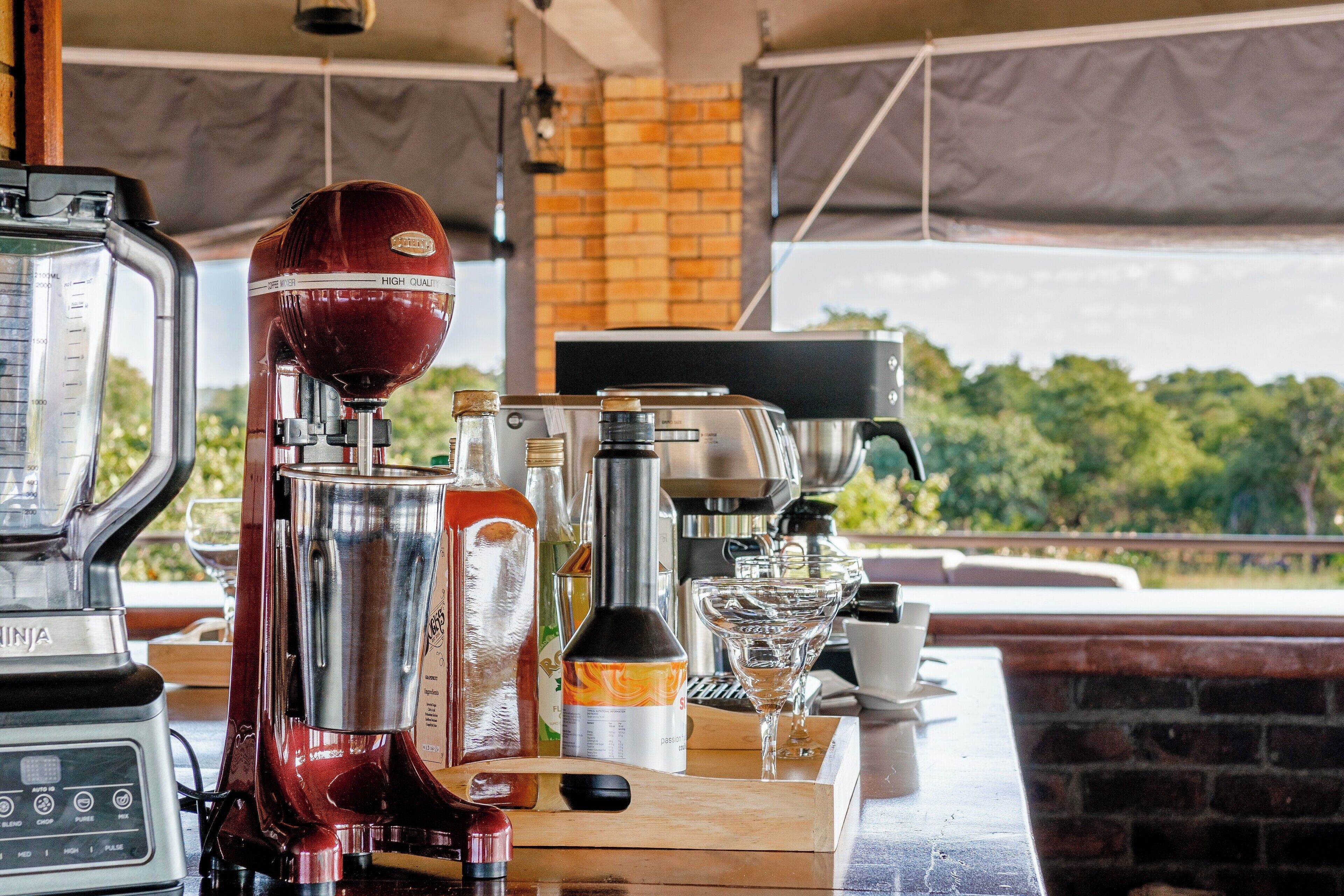 Uma área de bar com máquina de café, liquidificador e diversos frascos, com vista para o verde lá fora.