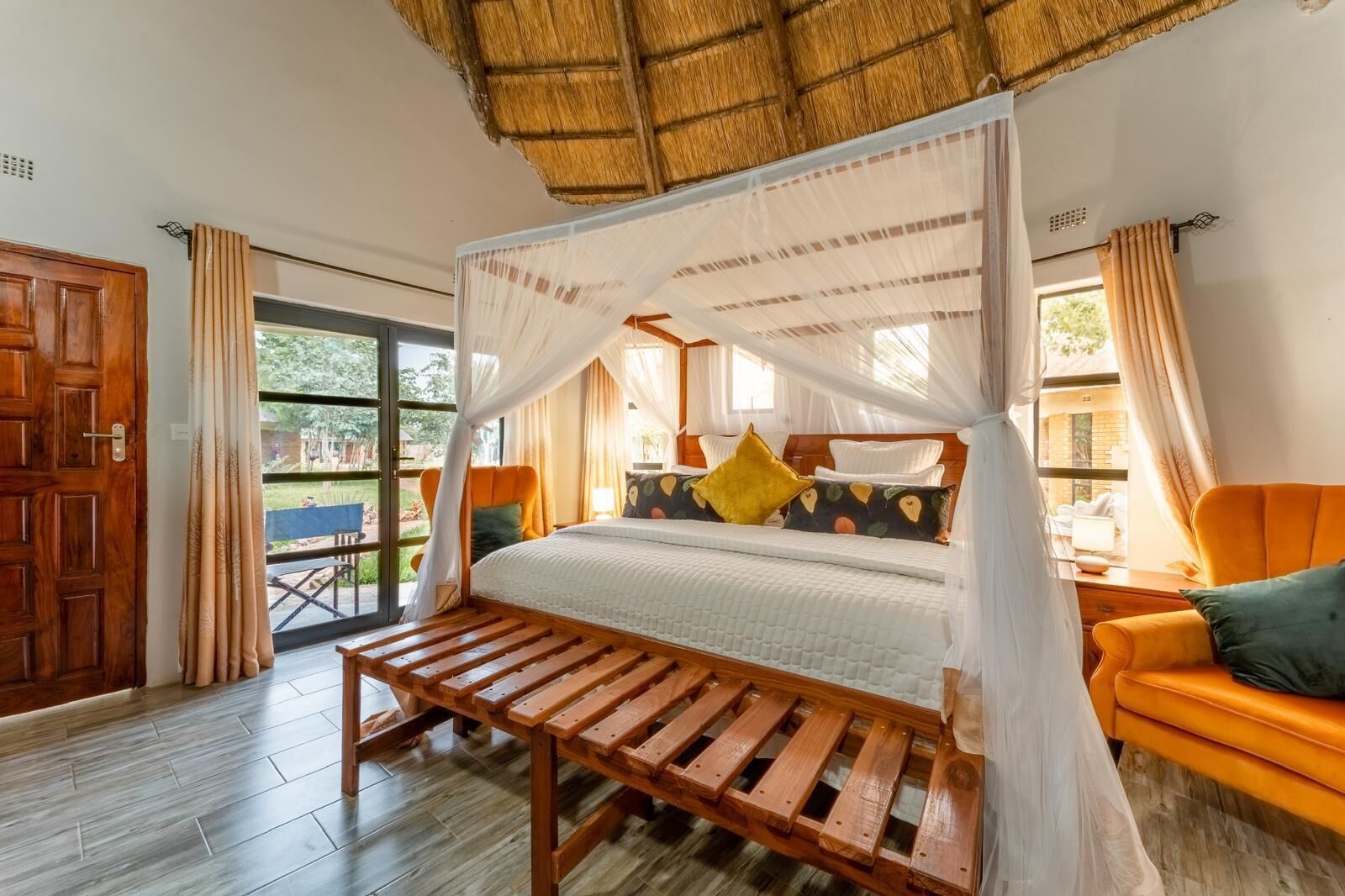 A room with a wooden bed, a bench, and a chair. There is a door and a window with curtains.