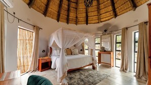 A bedroom with a canopy bed, wooden furniture, a thatched roof, and large windows.
