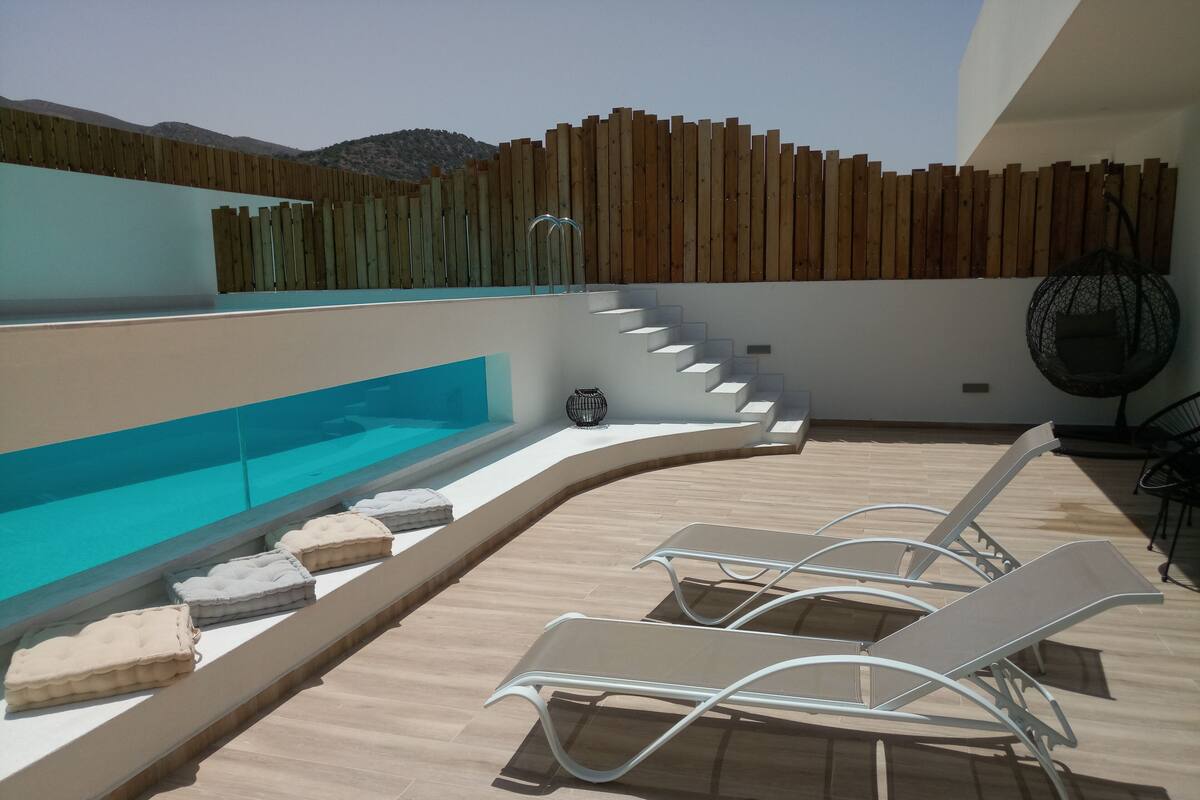A rooftop pool area with a wooden fence, a glass balustrade, and a seating area with lounge chairs.