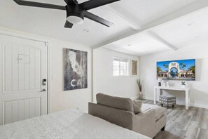 A bedroom with a bed, a ceiling fan, a painting, a desk, and a window.