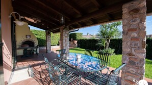 Un patio exterior con una mesa redonda de vidrio y sillas de metal, rodeado de pilares de piedra y un muro de piedra.