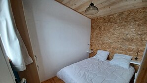 A bedroom with a wooden floor, a white bed with a quilted cover, a wooden headboard, and a hanging light fixture. - Tiny House in Méaudre in the heart of nature (Vercors plateau) (Autrans-Méaudre en Vercors)
