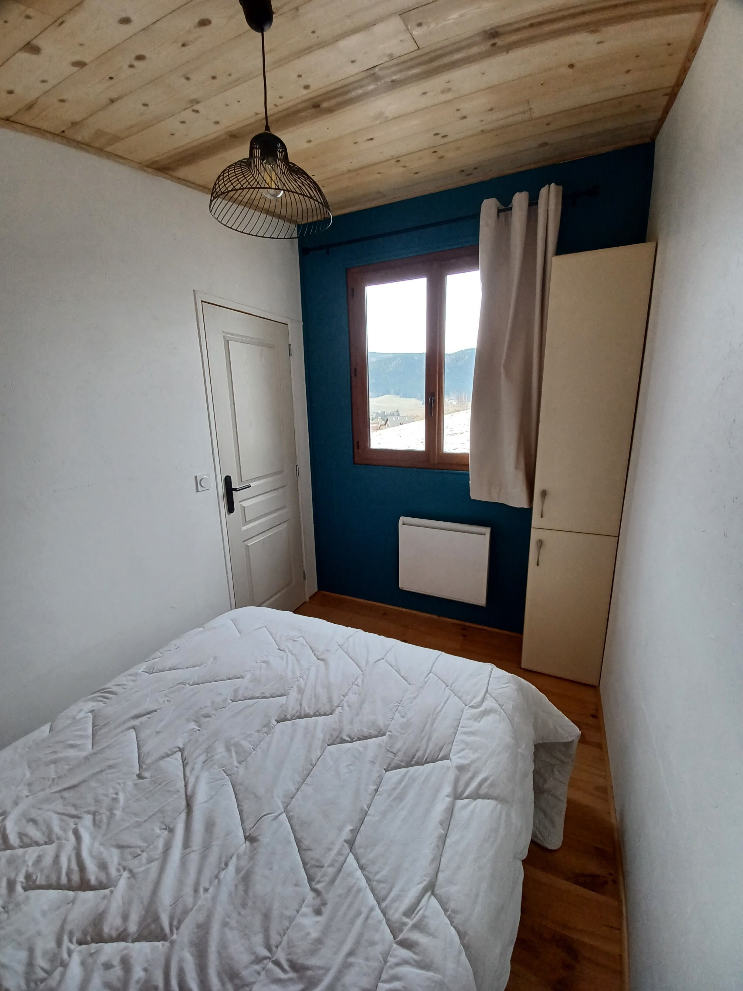 A bedroom with a wooden ceiling, a bed with a white quilt, a window with curtains, and a wardrobe.