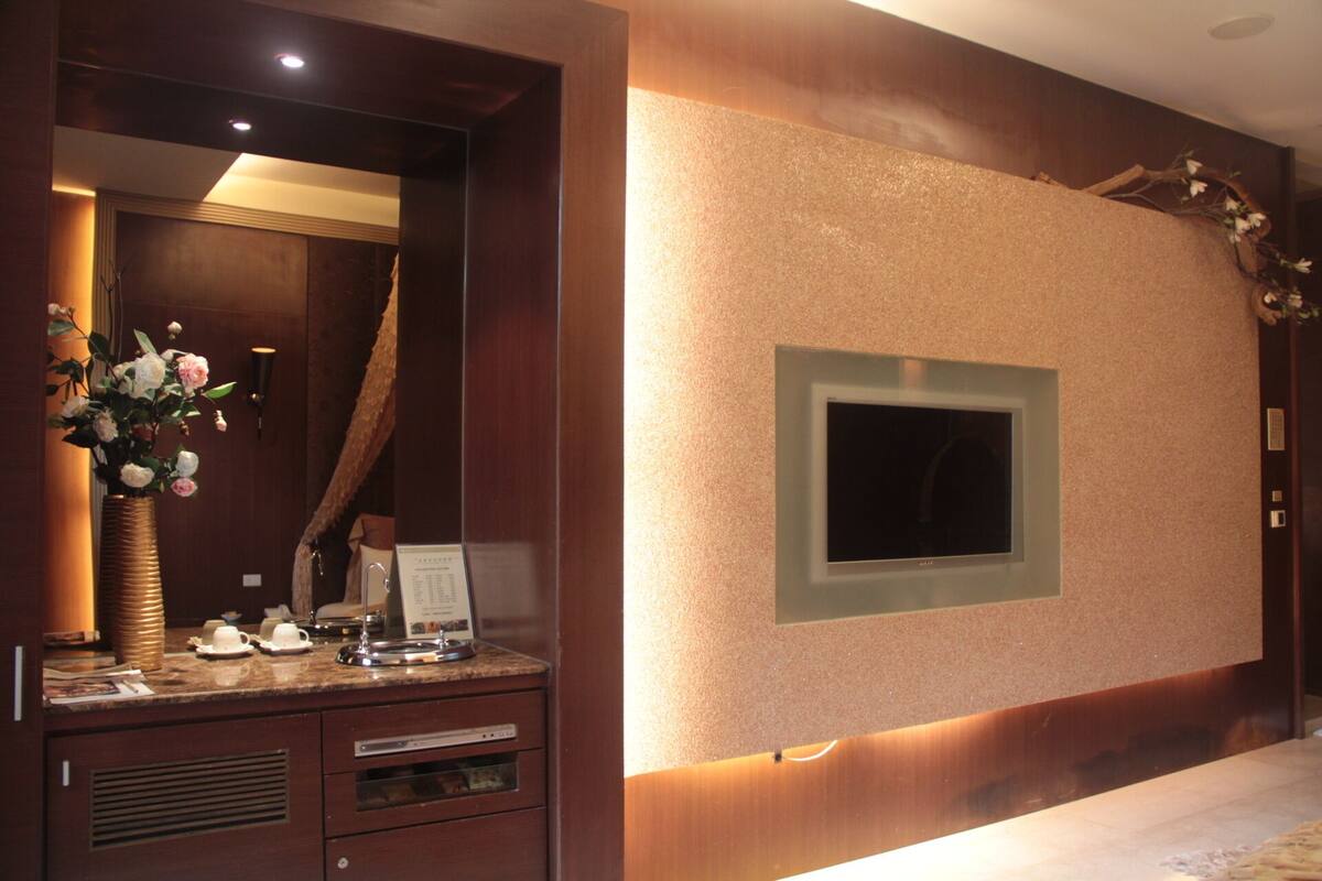 Habitación de hotel con un espejo grande, una tocador con un florero y flores, un televisor de pantalla plana montado en la pared y un mueble de madera con cajones.