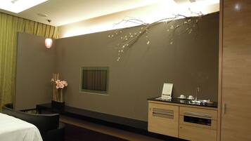 A hotel room with a bed, a small seating area, a vanity with a mirror, and a wall-mounted shelf.